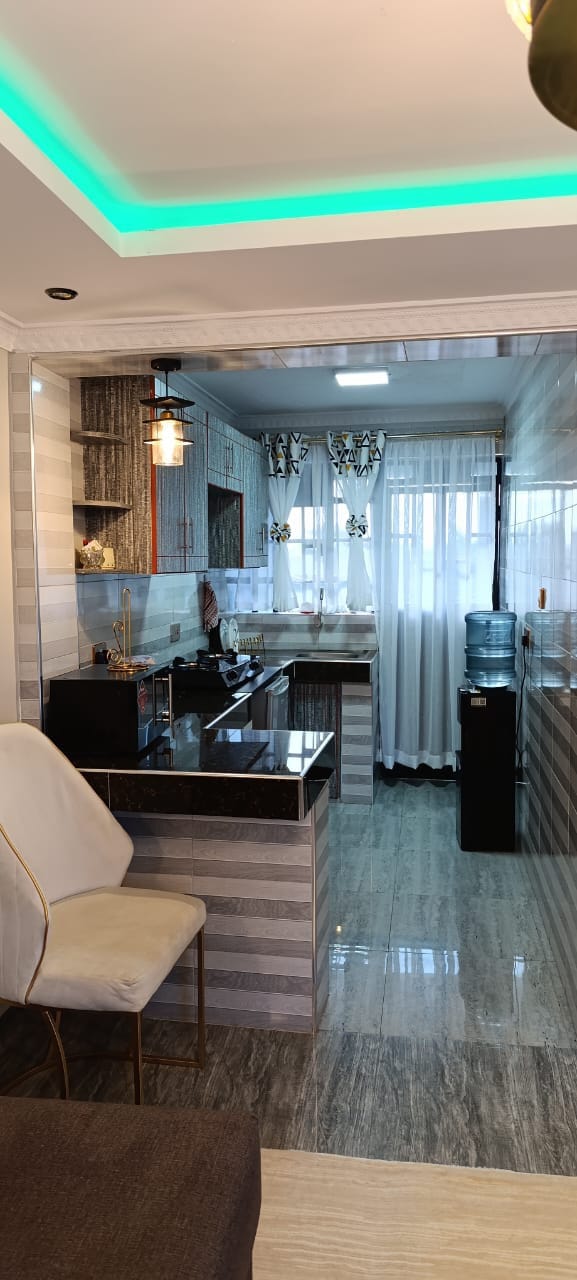 A modern kitchen is featured with dark countertops and light-colored cabinetry. Natural light enters through sheer curtains, illuminating the space. A water dispenser and a sleek design chair complement the kitchen's contemporary style, creating a functional and inviting atmosphere.
