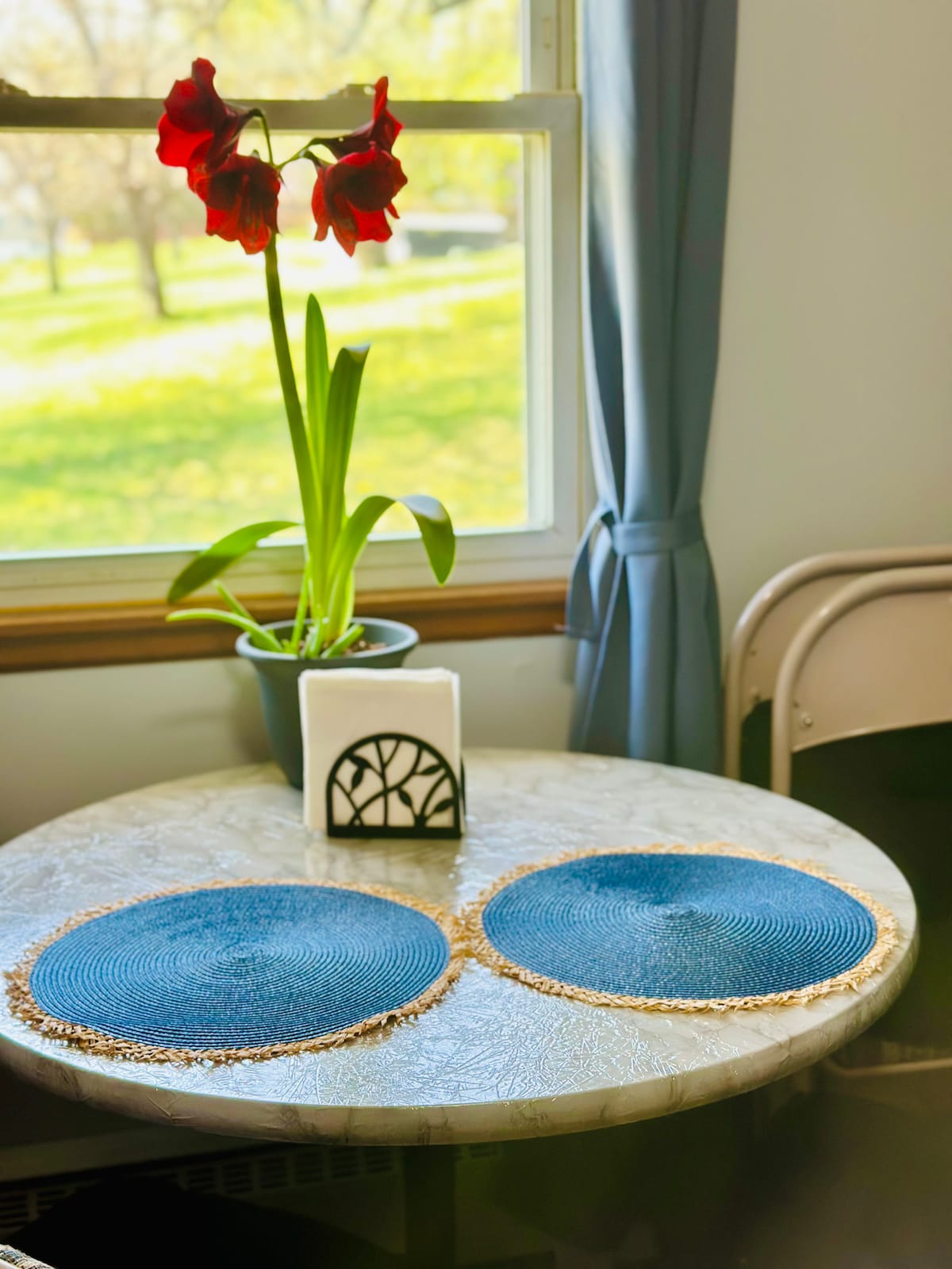 A small, round table is set by a window, featuring two woven place mats. A potted plant with vibrant red flowers sits in the center, complemented by a simple candle holder. Natural light floods the area, highlighting the greenery outside.