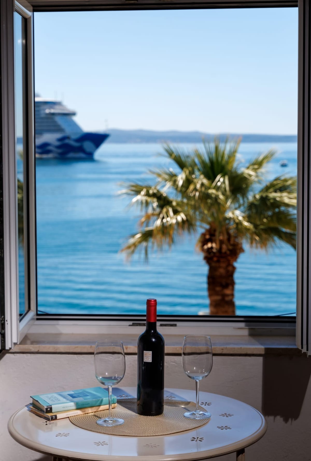 A table set by the window features a bottle of wine and two glasses, inviting relaxation. The view showcases a calm sea, with palm trees swaying gently in the foreground and a cruise ship visible in the distance.