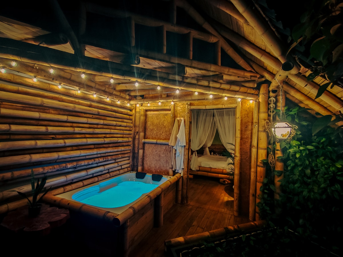 An intimate space features a small, illuminated pool surrounded by bamboo walls. Soft lighting enhances the cozy atmosphere, while a comfortable bed is seen in the background, draped with sheer curtains. Plants are positioned around the interior, adding a touch of nature.