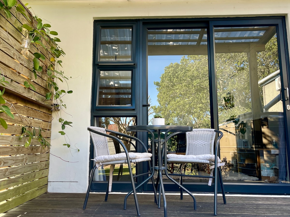 A private deck is showcased with a small table and two chairs, offering a comfortable outdoor seating area. Sunlight filters through the glass doors, illuminating the space and reflecting the greenery from the surrounding foliage.