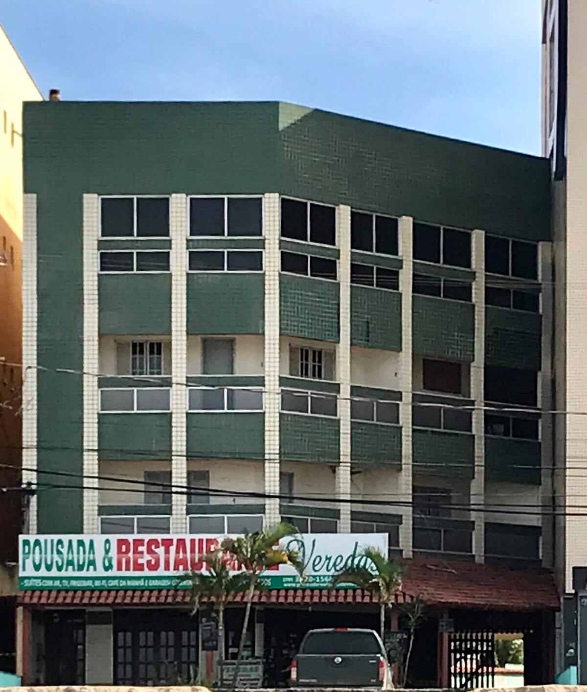 A three-story building features a distinct green tiled façade and multiple windows on each level, allowing natural light to fill the interior. The ground level includes a signage for a restaurant and guesthouse, indicating a lively atmosphere at the location.