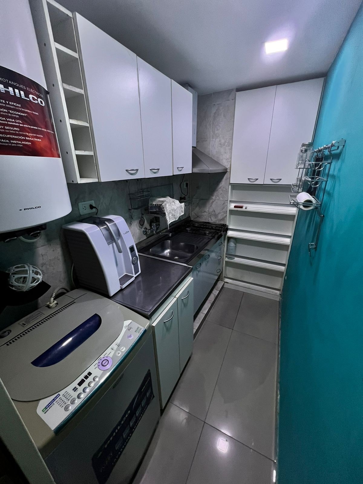A compact kitchen area is shown, featuring modern appliances including a washing machine and a small refrigerator. Cabinets above provide storage, while a countertop holds an electric stove and workspace. Light gray tiles cover the floor, and a vibrant blue wall offers a pop of color.
