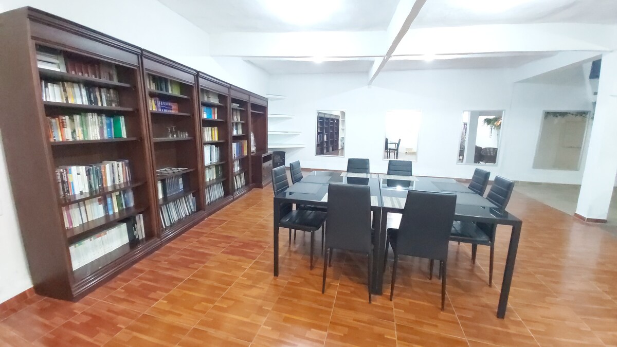 A spacious communal area is shown, featuring a large black table surrounded by eight black chairs. A wooden bookshelf filled with books occupies one wall, while mirrors reflect the room's layout. The floor is tiled in warm tones, providing an inviting atmosphere.