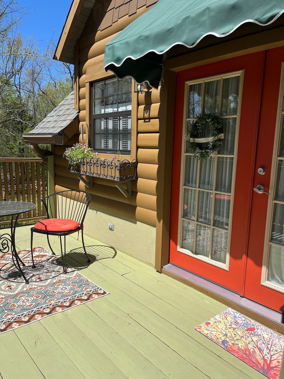 A welcoming deck area is shown, featuring a round metal table with two cushioned chairs. Colorful area rugs are placed beneath the table and doorway. Potted plants are arranged on the railing, and a decorative wreath adorns the door, framed by large glass panels.