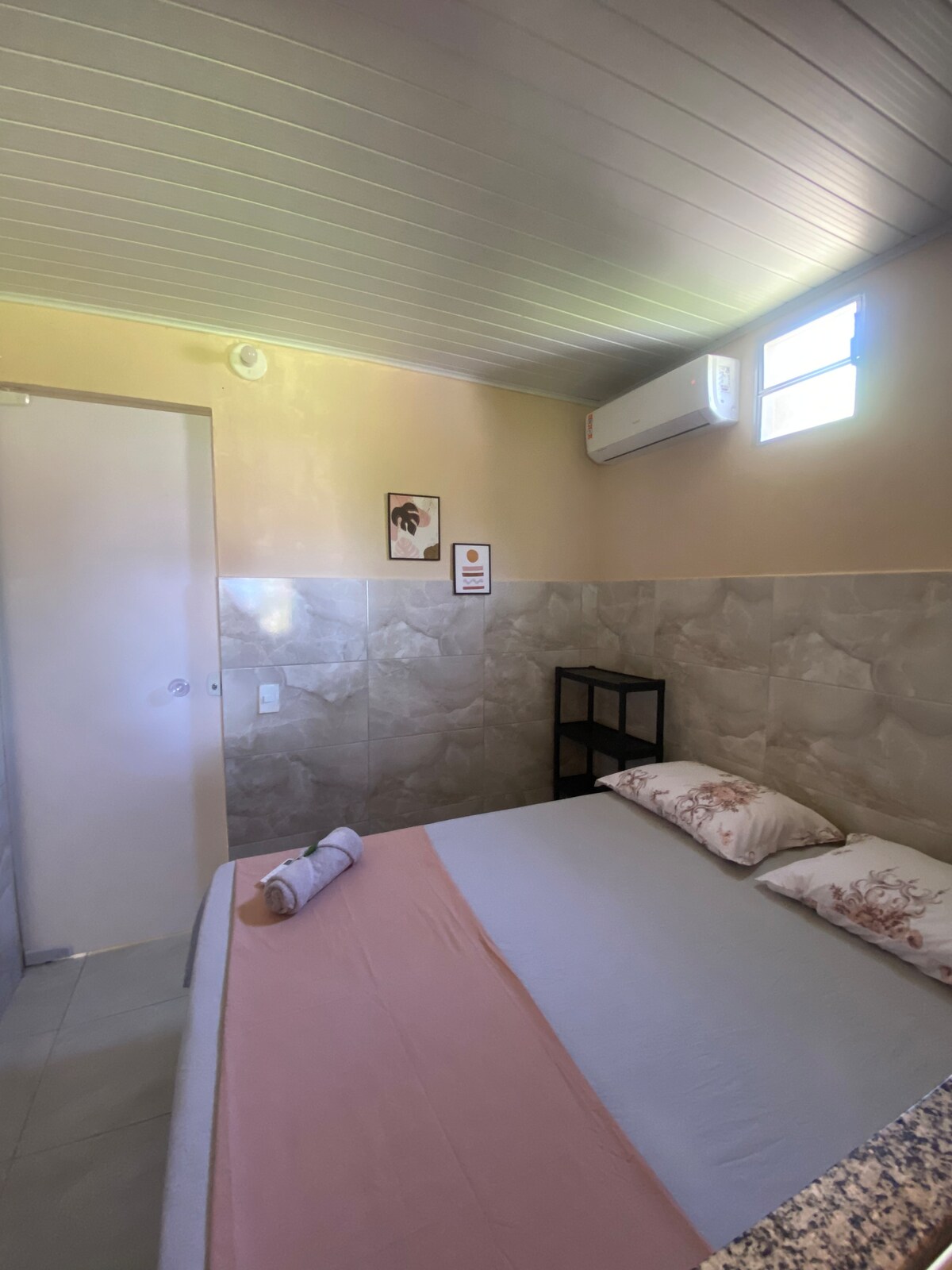 A simple bedroom is depicted, featuring a spacious bed with neutral bedding and accent pillows. A small black shelving unit stands against a light-colored wall, and an air conditioning unit is installed near the ceiling. Natural light enters through two small windows.