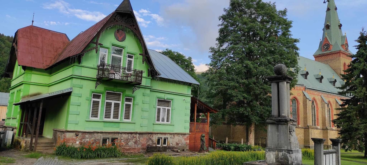 A charming green house is positioned alongside a church with a tall steeple and decorative roof. Lush greenery surrounds the property, complemented by well-maintained landscaping. A stone pathway leads to the entrance, offering easy access to the inviting exterior.