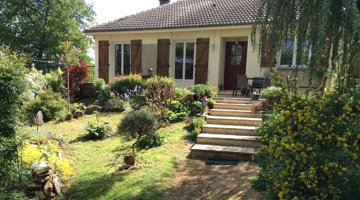 A charming entryway is accentuated by a set of wooden steps leading up to the house. Lush greenery and colorful flowers, including yellow blooms, surround the pathway. A small seating area with a table and chairs is situated to the right, inviting relaxation in the garden space.