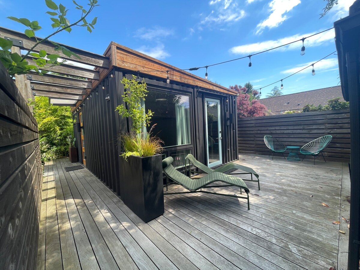A modern deck area is surrounded by wooden fencing, featuring two lounge chairs and a tall planter with greenery. The container home is visible with large glass doors, allowing natural light into the space. String lights are hung above, adding a warm ambiance.