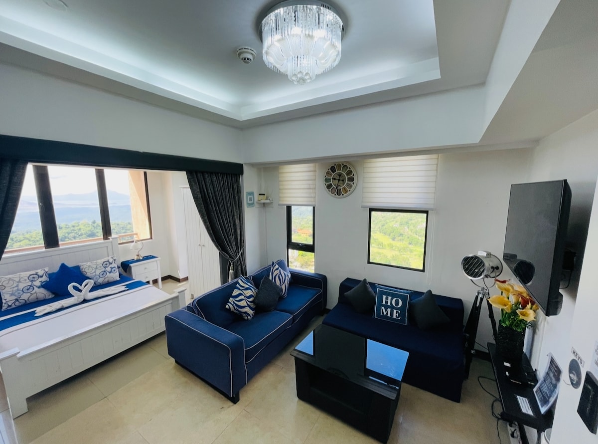 A modern living area showcases a blue sofa with accent pillows and a glass coffee table. Natural light filters through the large windows, framing scenic views. Adjacent, a cozy daybed with decorative cushions adds a touch of comfort. A statement chandelier is suspended from the ceiling.