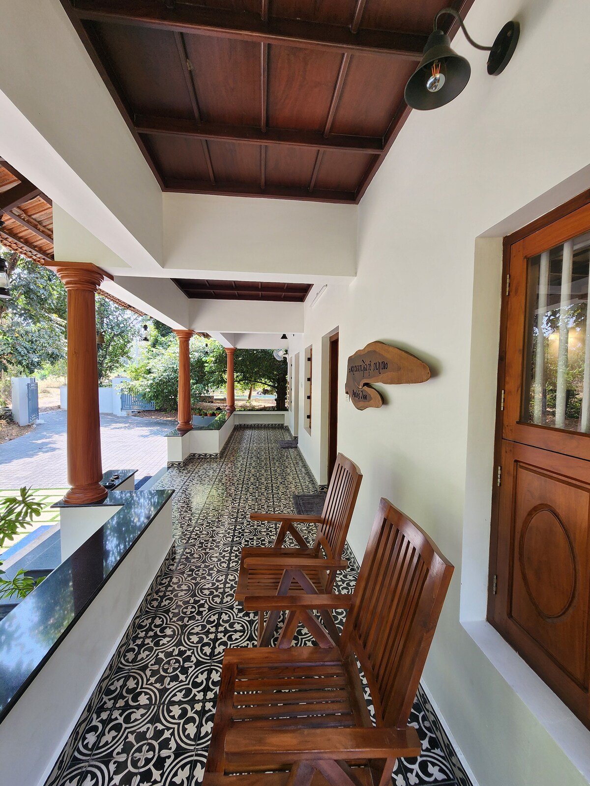 A covered outdoor seating area is presented with two wooden chairs positioned against a decorative tiled floor. Sunlight filters through the trees, providing a natural setting by the path, while light fixtures hang from the wooden ceiling, emphasizing the traditional architectural details.