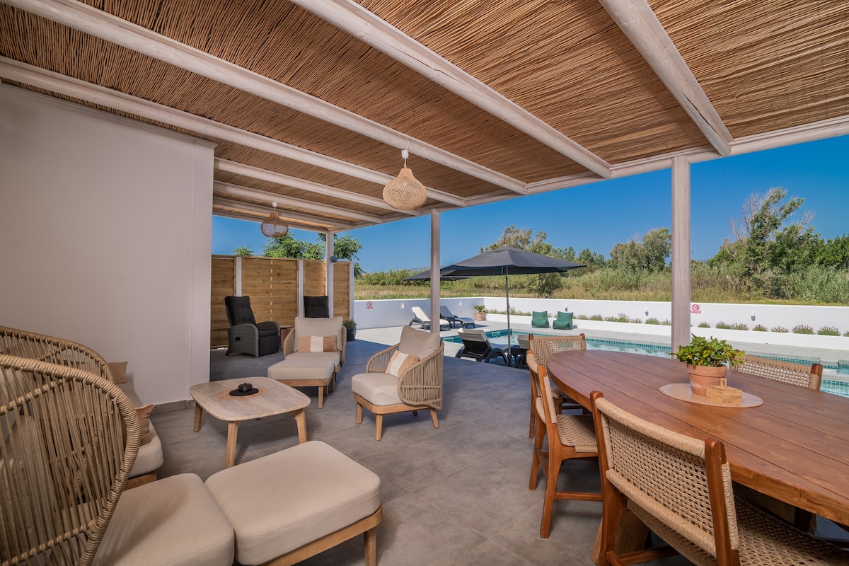 An outdoor living space is presented with various seating options, including woven chairs and a large dining table. Natural light illuminates the area, while shades from umbrellas provide comfort. A view of the private pool and surrounding greenery is visible in the background.