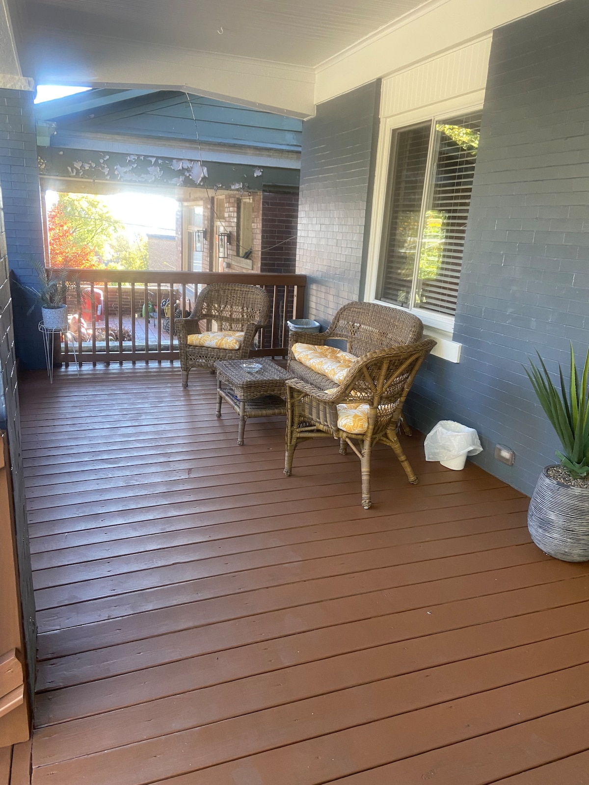 A front porch is presented with a wooden floor and natural light streaming in. Two wicker chairs are paired with a small table, all featuring soft yellow cushions. A potted plant adds a touch of greenery, enhancing the relaxed outdoor space.