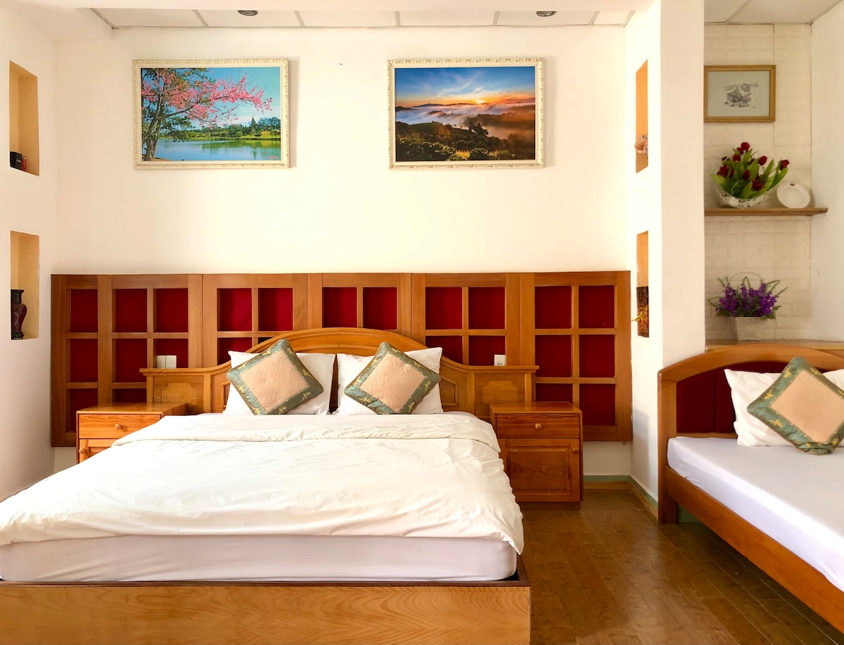 A well-furnished room features a spacious king bed and a single bed. Wooden furniture adds warmth, complemented by red and neutral wall decor. Artwork depicting landscapes adorns the walls, while natural light fills the space through unobstructed windows.