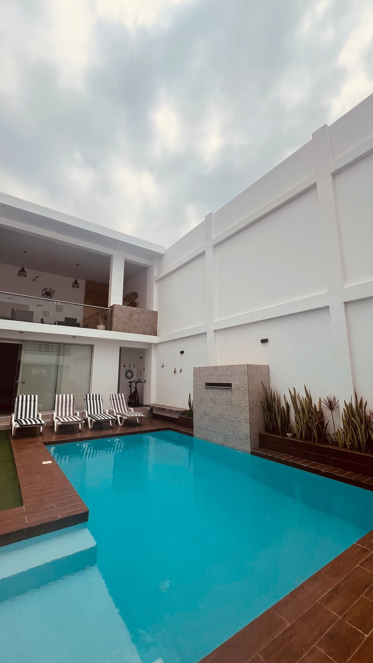 A serene pool area features clear blue water reflecting the sky, framed by a wooden deck. Four striped lounge chairs are positioned alongside the pool, with elegant landscaping including tall plants enhancing the space. The modern architecture is complemented by spacious walls in soft neutral tones.