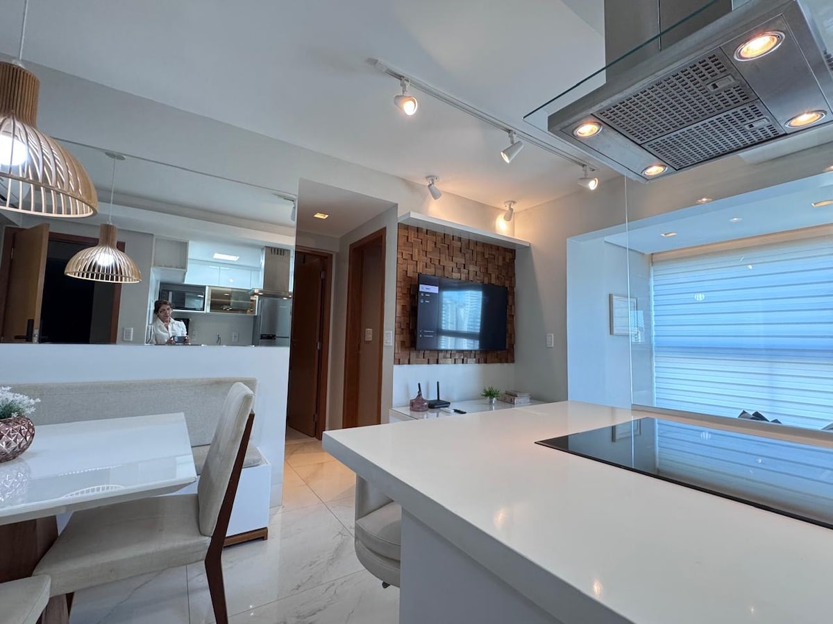 A modern kitchen and dining area are featured with white cabinetry and sleek countertops. A dining table with light-colored chairs accommodates multiple guests, while a flat-screen TV is mounted on a wooden accent wall. Natural light filters through a nearby window with soft blinds.