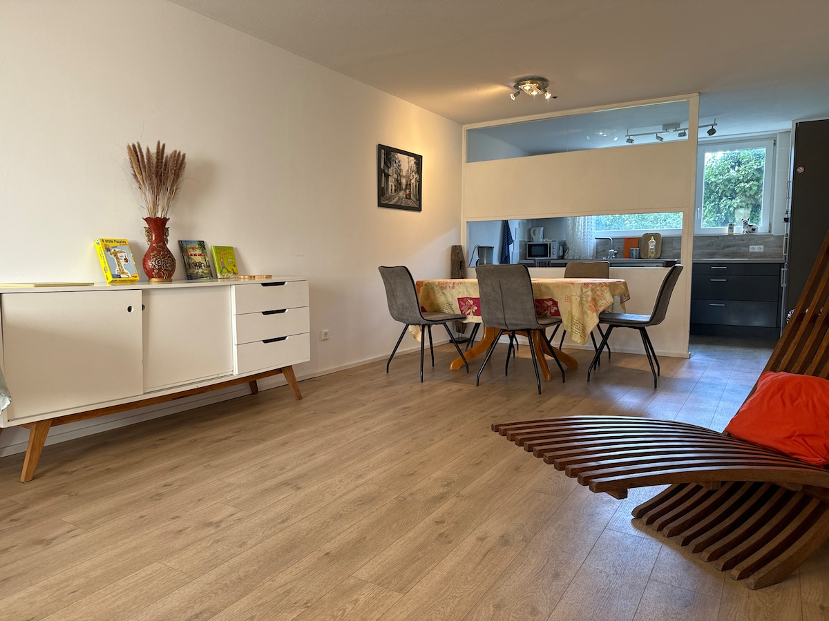 A spacious living area features light wood flooring and clean white walls. A dining table with four upholstered chairs sits adjacent to a sleek kitchen, while a decorative cabinet holds framed photos and books. A wooden lounge chair adds a unique touch to the room.