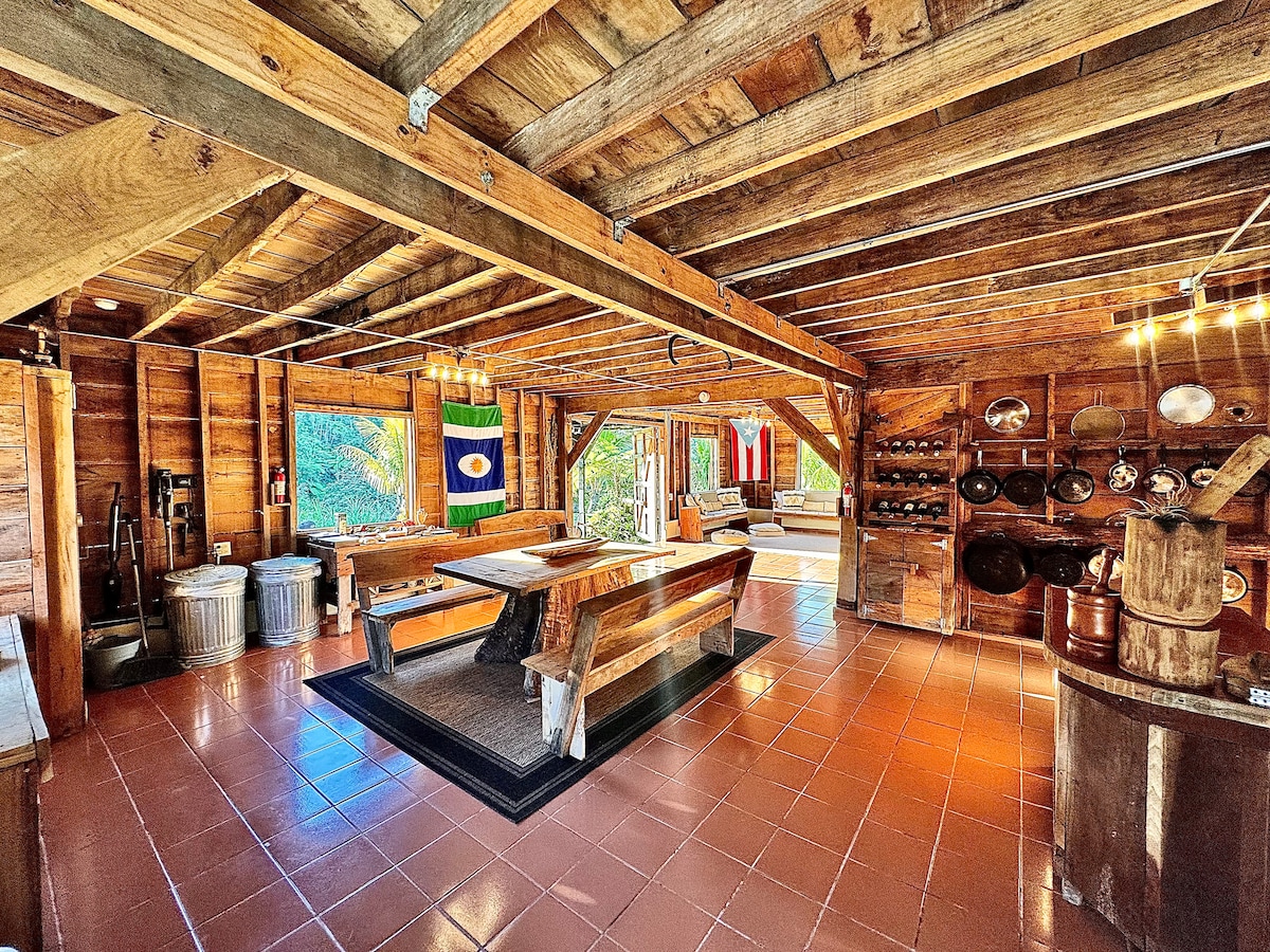 An open-concept living area features rustic wooden beams and warm terracotta tiles. A large dining table is set at the center, surrounded by benches. Shelves display handcrafted kitchenware, while vibrant flags add a touch of local culture. Natural light filters in through large windows, revealing outdoor views.