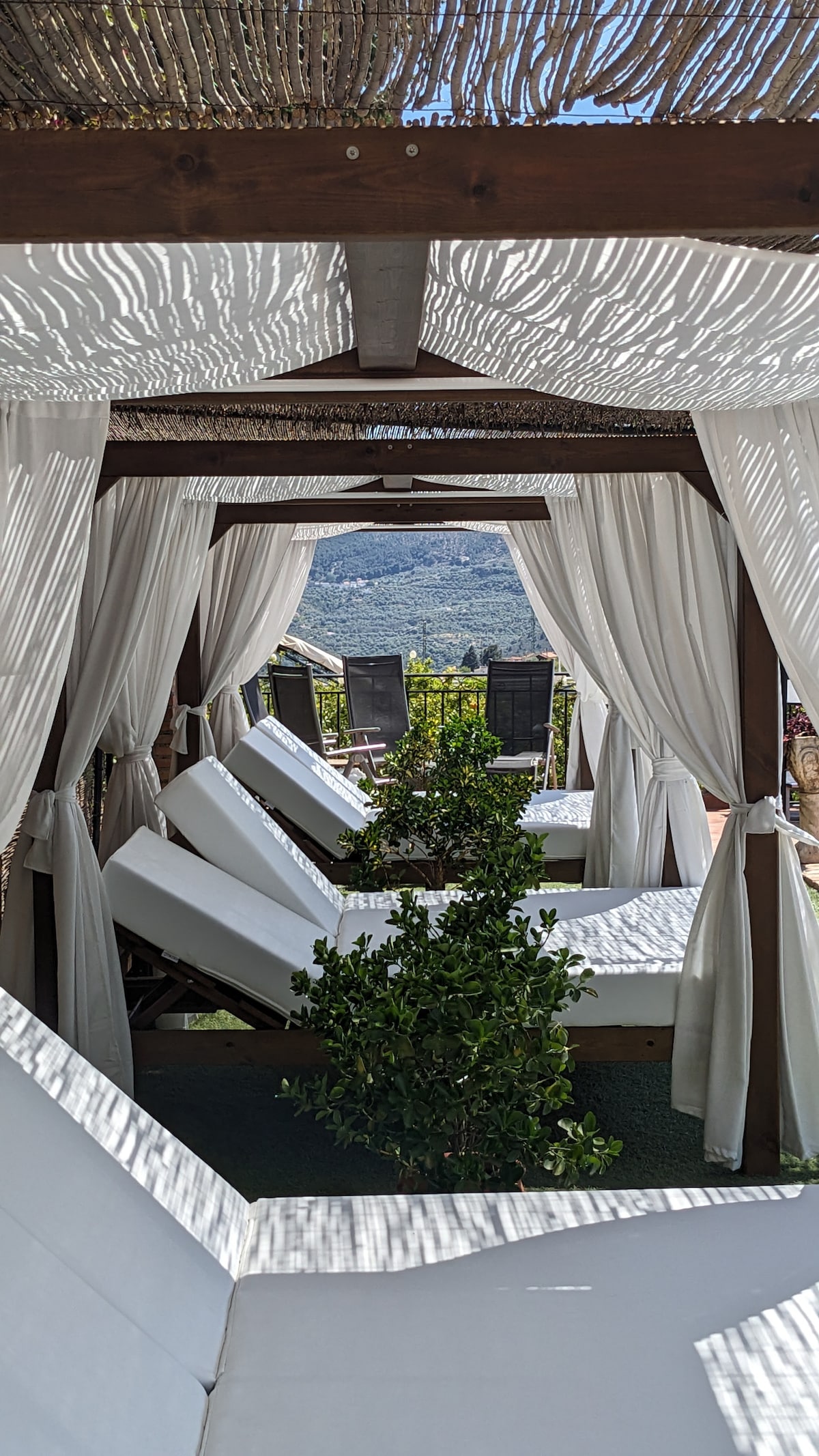 A shaded lounging area features white, reclining chairs positioned under a canopy. Light filters through the fabric, creating gentle patterns. Lush greenery is placed between the chairs, with scenic views of rolling hills visible in the background.
