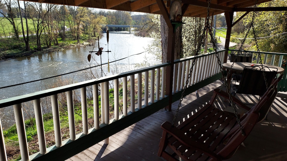 A shaded outdoor area features a wooden swing, suspended above a deck overlooking a serene creek. The gentle flow of water can be seen, framed by trees and grass along the banks, creating a peaceful setting for relaxation.