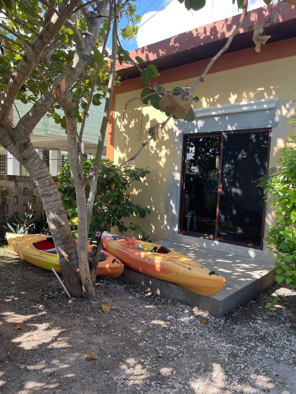 Two brightly colored kayaks, one yellow and one orange, are positioned by a concrete patio area. A nearby tree provides partial shade, while the entrance to the suite is visible through large glass doors, opening to a quiet outdoor space with surrounding greenery.