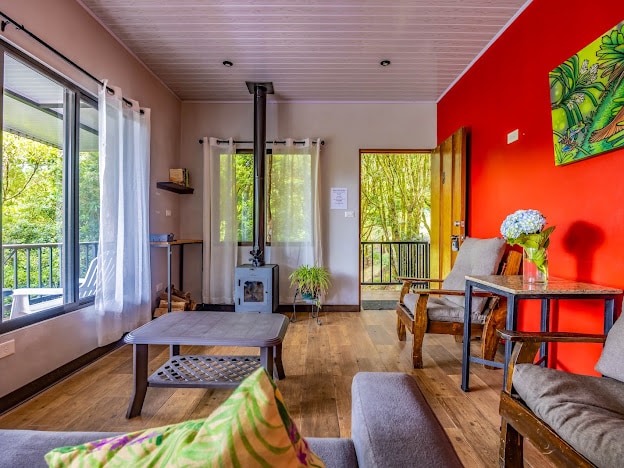 A cozy living area features a wood stove against a vibrant red accent wall. Large windows offer forest views, with natural light illuminating the space. Comfortable seating arrangements include two armchairs and a sectional sofa, complemented by a wooden coffee table and a decorative flower arrangement.