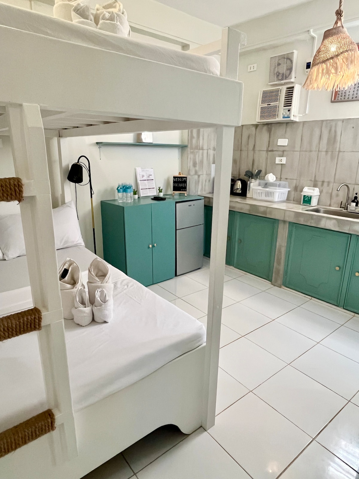 The room features two double deck beds, accommodating up to six guests. A small kitchenette is equipped with a refrigerator and cooking utensils. Natural light illuminates the clean, tiled flooring, while a cozy ambiance is created by a woven pendant light.