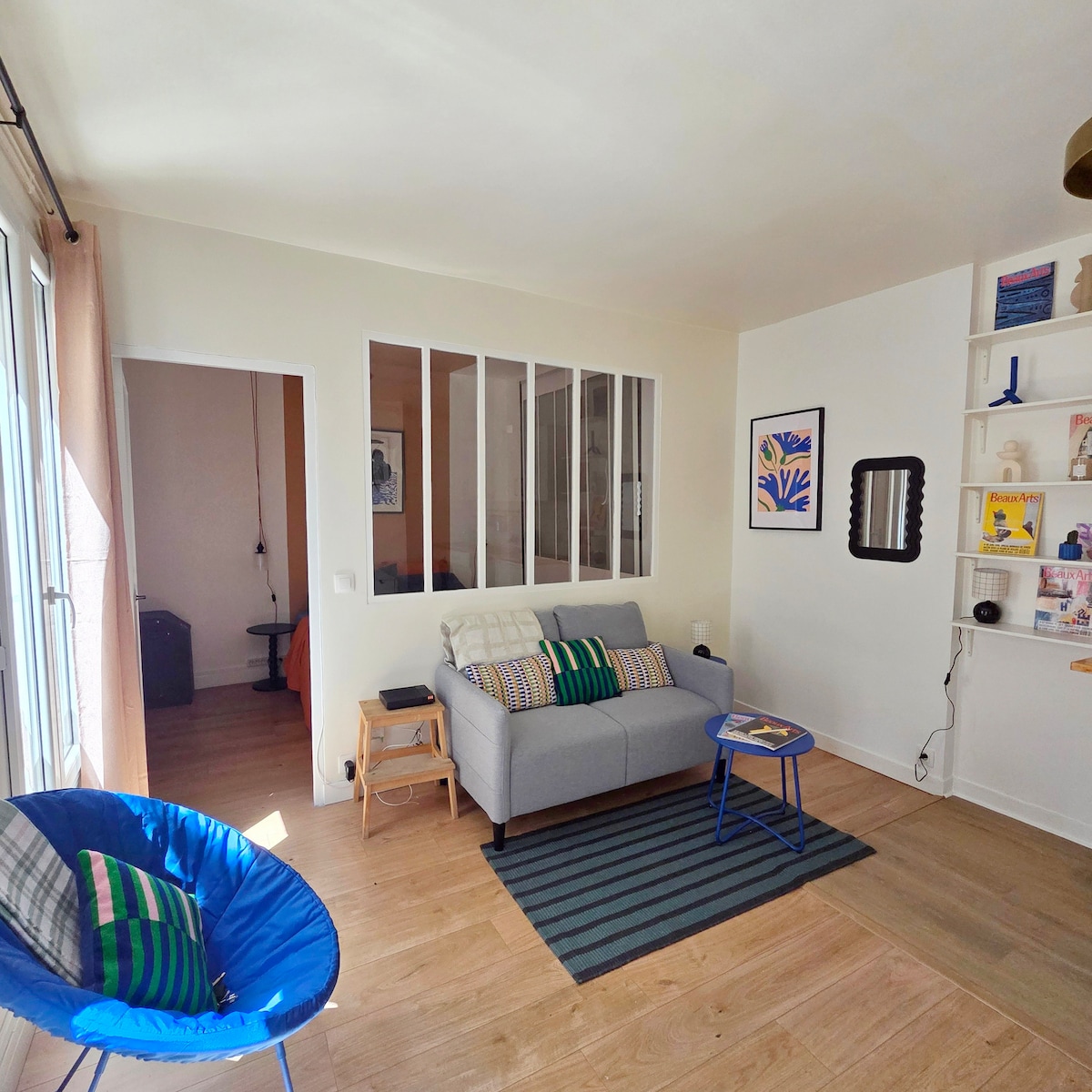A light-filled living area features a comfortable gray sofa adorned with colorful throw pillows. A small blue table is positioned next to it on a striped rug. An accent chair adds a pop of color, and a glass partition separates the living area from the bedroom.