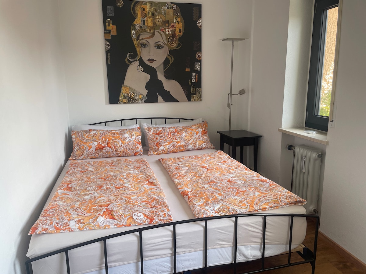 A well-designed bedroom features a modern black bed frame with patterned bedding in warm hues. A bedside table is positioned next to the bed, while a bright piece of wall art adds a touch of color. Natural light streams in through the nearby window.