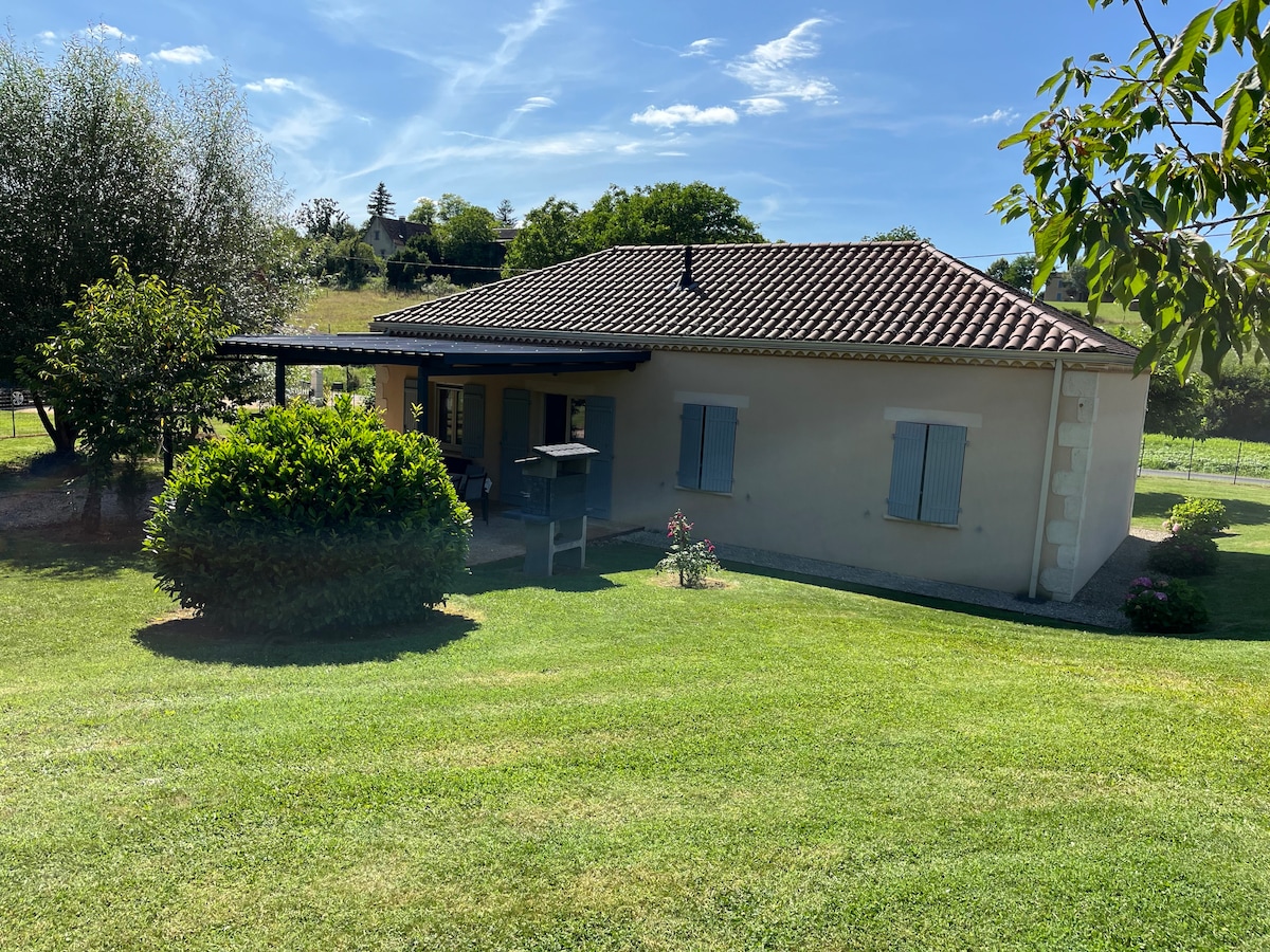 A single-story house is set against a backdrop of lush greenery and blue skies. The property features a garden with well-kept grass, flowering plants, and trees. A shaded terrace extends from the house, providing inviting outdoor space for relaxation.