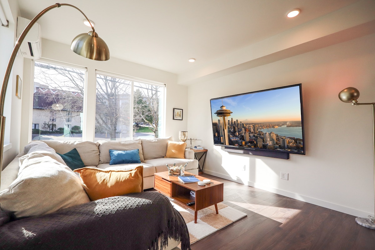 The living room features a modern sectional sofa adorned with colorful throw pillows. A large TV displays a cityscape image on the wall, complemented by oversized windows that allow natural light to brighten the space.
