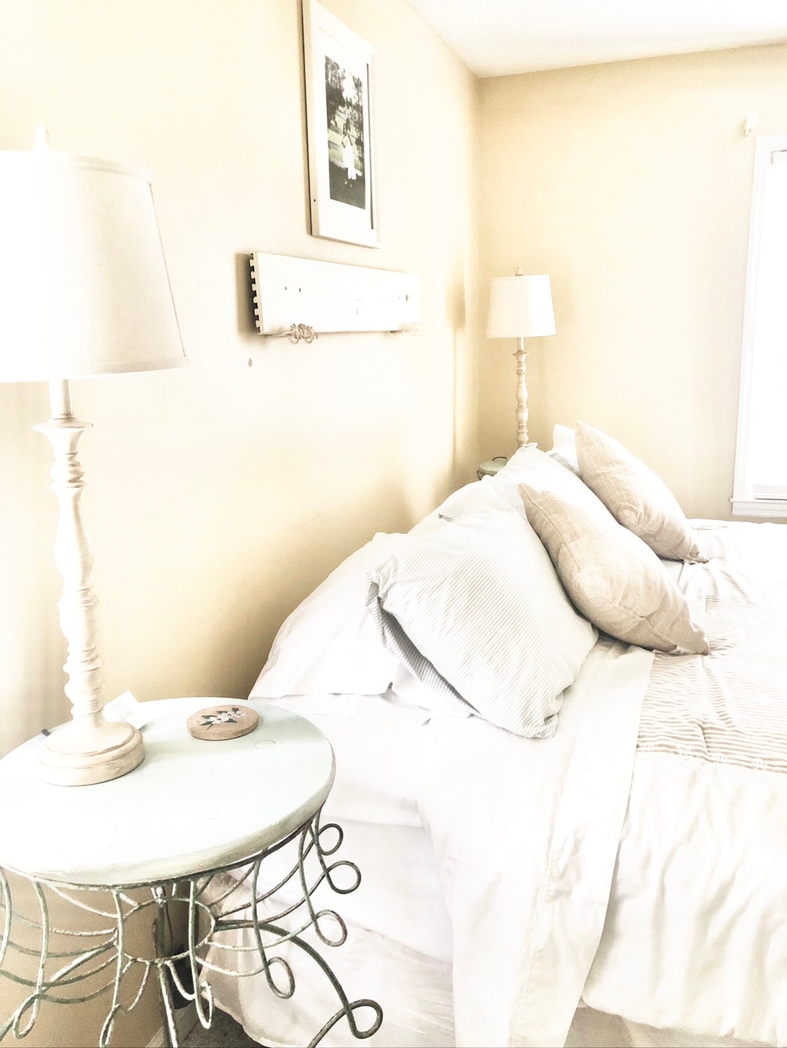 A well-appointed bedroom features soft beige walls and natural light streaming in through a window. The spacious bed is adorned with light-colored linens and decorative pillows. Beside the bed, a quaint side table holds a lamp and a small decorative item.