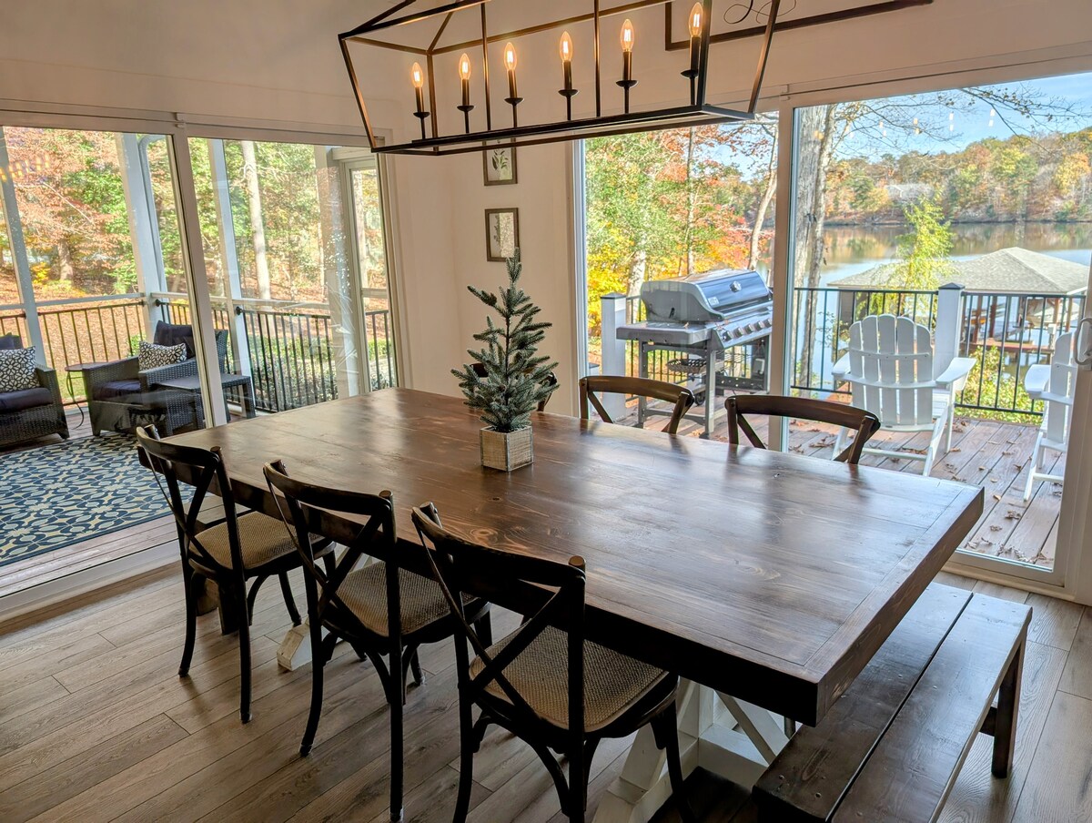 A long wooden dining table with six chairs is positioned beneath an artistic light fixture. Large glass doors provide a view of the lake and outdoor seating, while natural light floods the space, creating a welcoming dining area.