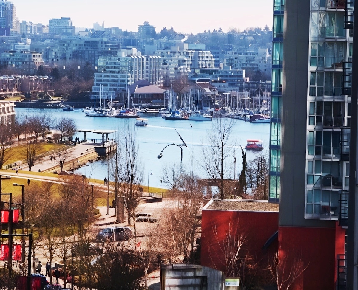 A scenic view of the waterfront features moored yachts and boats gently bobbing on the water. Modern buildings line the shore, framed by bare trees. The vibrant green park offers pathways and benches for leisurely enjoyment along the water's edge.