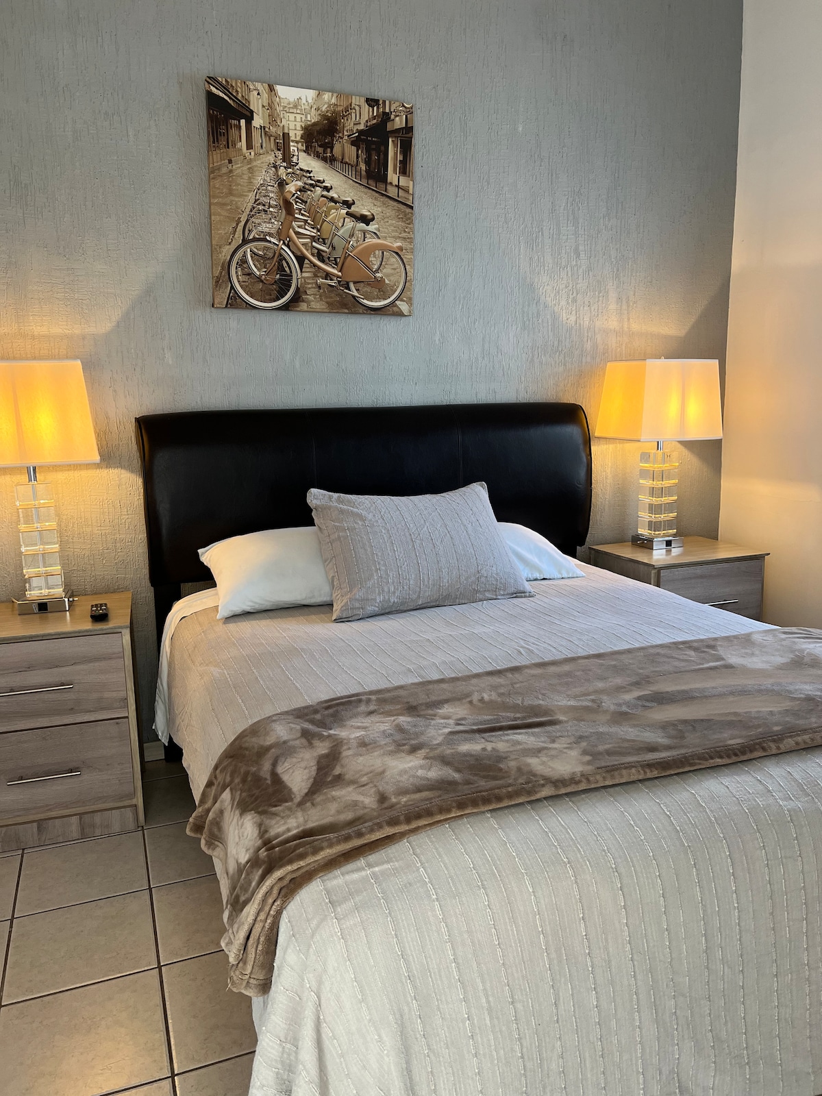 A neatly arranged sleeping area features a dark headboard with a soft white bedspread, complemented by decorative pillows. Flanking the bed are two bedside tables, each topped with elegant lamps providing gentle illumination. A framed image of bicycles decorates the wall above the bed.