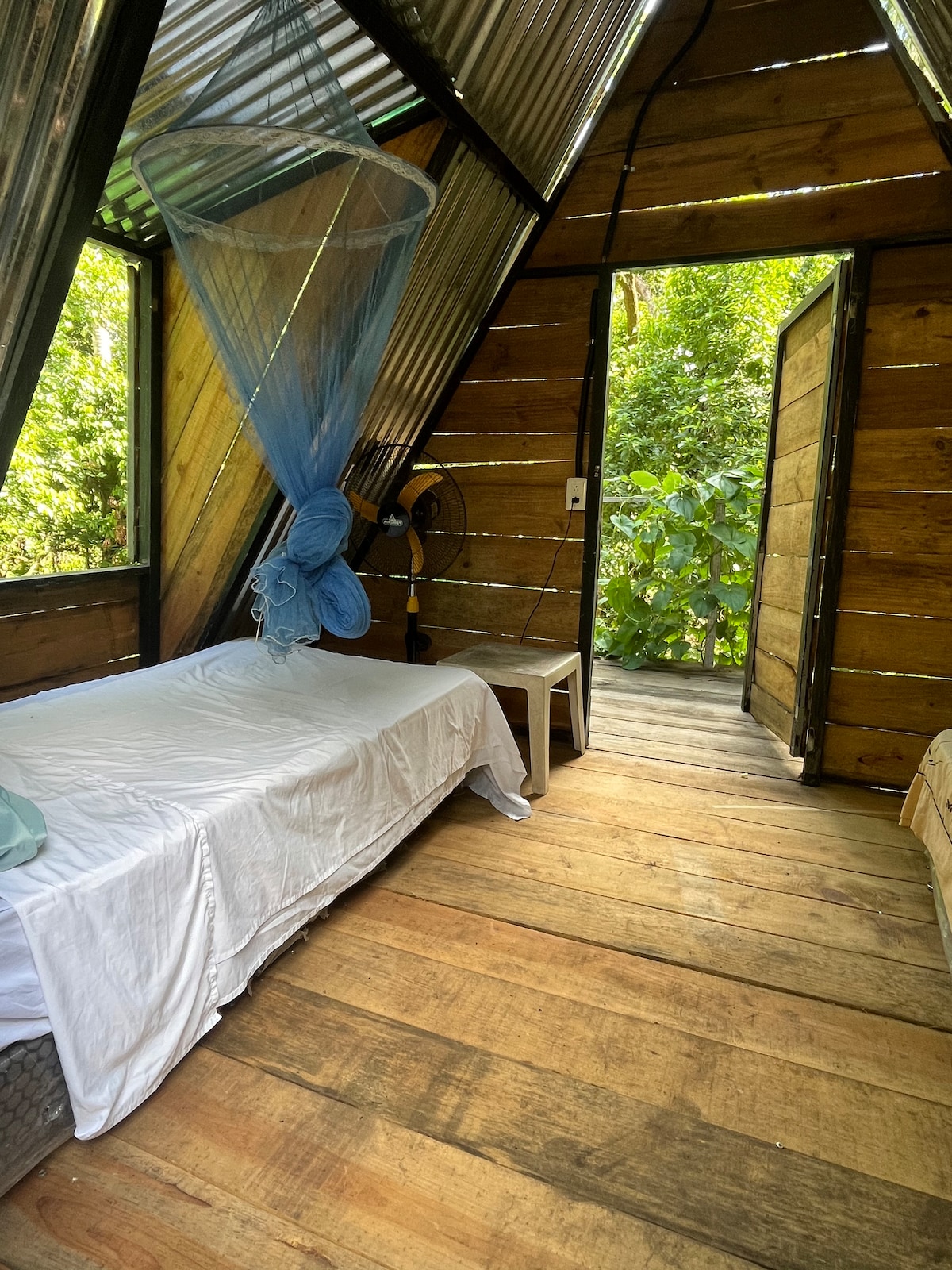 A simple sleeping area features a bed with a white coverlet and a mosquito net overhead. Natural light filters in through an open door, revealing lush greenery outside. A small white chair and a fan are positioned within the space, enhancing comfort.