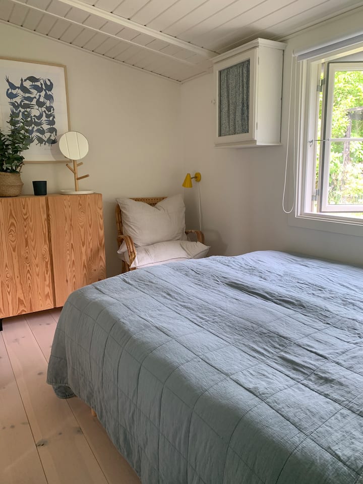 Bright and cozy bedroom with garden view. Here you will find a 140cm bed, a practical wardrobe, a small shelving unit, and a charming wicker armchair with soft pillows - perfect for relaxing.