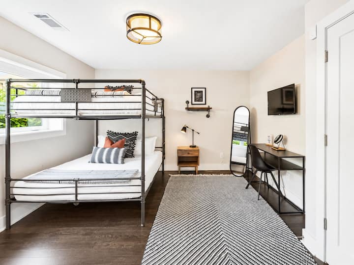 Metal bunk bed with striped bedding offers a queen over queen setup - Includes a wood nightstand, reading lamp, and minimalist desk with modern mirror - Bright window and geometric rug enhance the clean, contemporary vibe