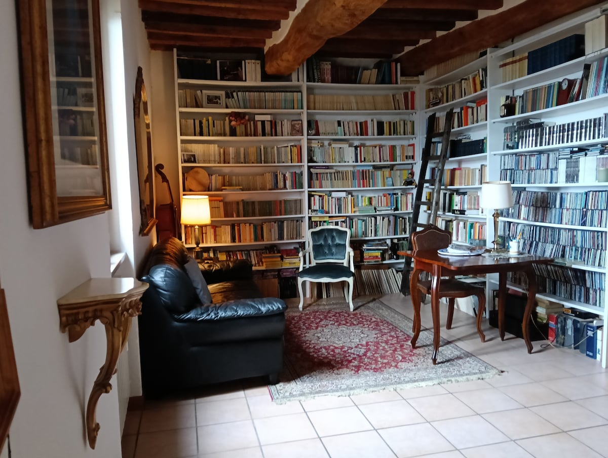 A charming reading area is filled with extensive bookshelves lined with books and CDs. A deep leather sofa complements a wooden desk and a light blue chair, while soft lighting creates a welcoming ambiance. A decorative rug enhances the cozy feel of the space.