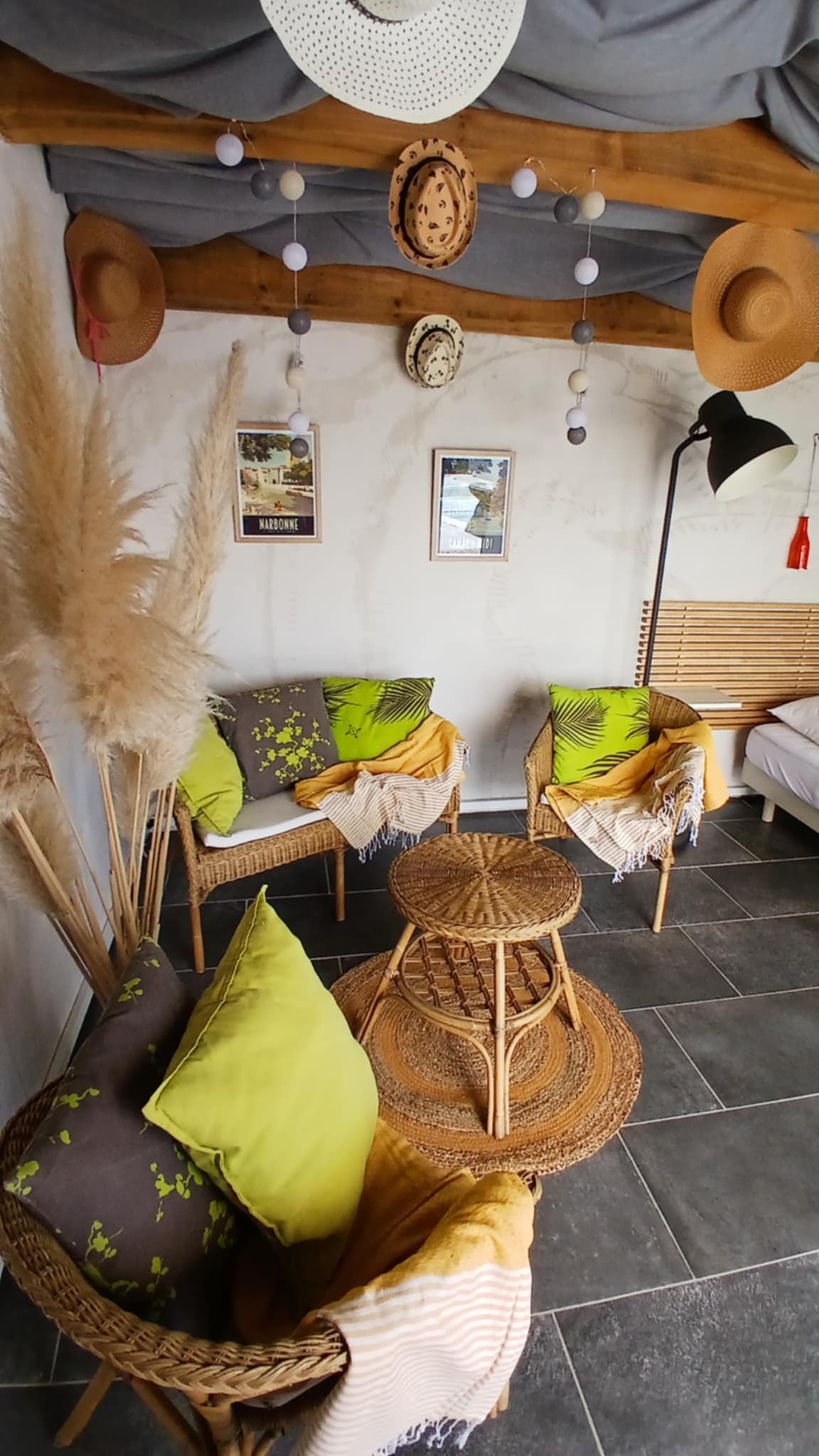 A cozy seating area is arranged with two woven chairs adorned with green and patterned cushions, accompanied by a matching circular table. Decorative hats hang from the ceiling beams, and framed artwork provides visual interest to the space with soft lighting illuminating the room.