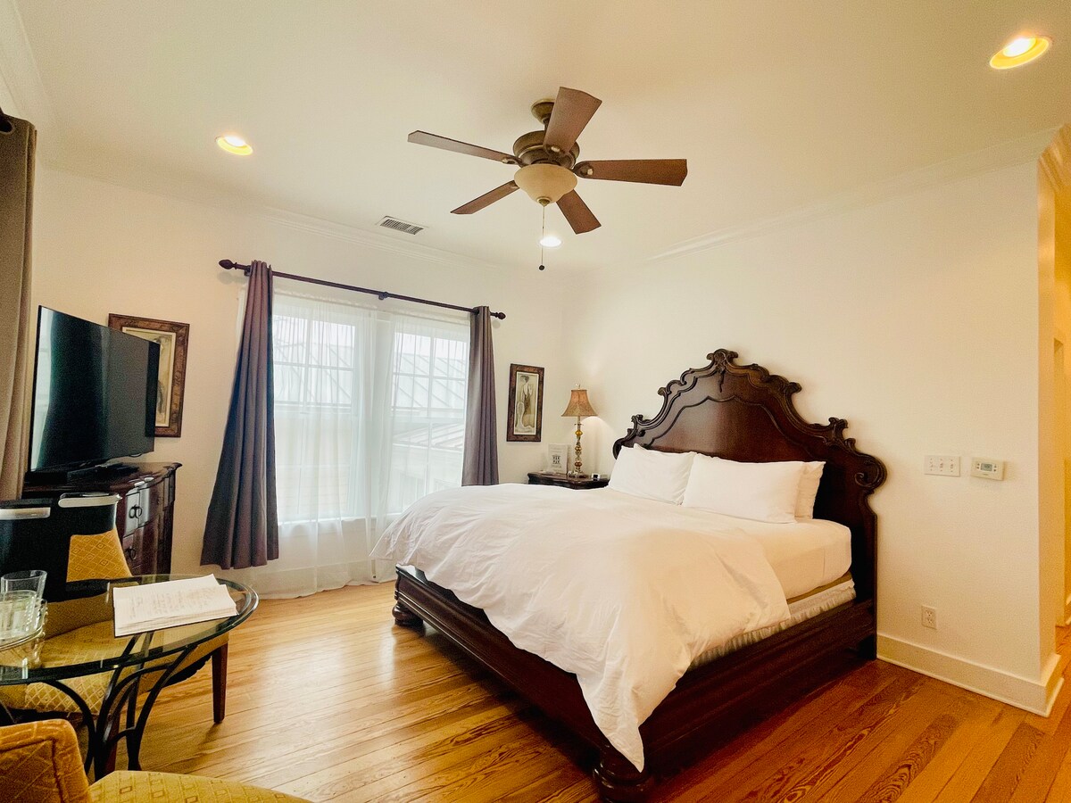 A spacious sleeping area features a large bed with crisp white bedding, complemented by a decorative wooden headboard. Natural light filters through sheer curtains, enhancing the warm wood flooring. A flat-screen television is positioned on a stand, while a cozy seating area invites relaxation.