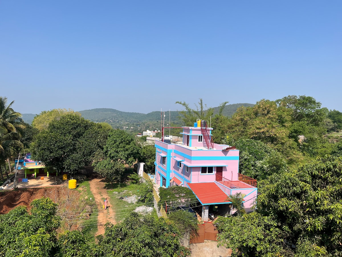A two-story home featuring a vibrant pink facade and a contrasting red roof is set amidst a lush landscape. Surrounding hills and trees create a serene backdrop. A playground area is visible nearby, complemented by a small yellow structure.