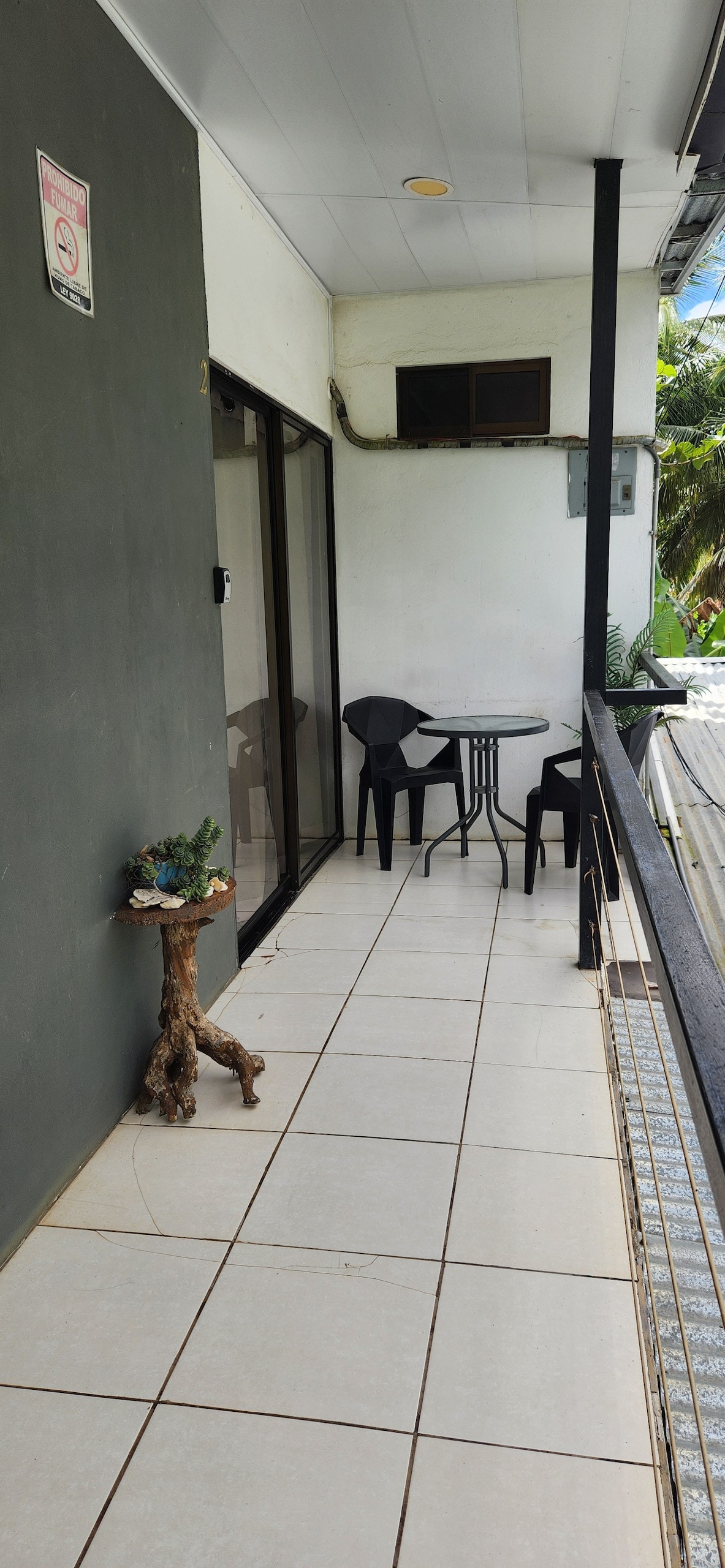A private balcony features a small round table and two black chairs, surrounded by tiled flooring. A plant decorates one side, while the entrance is marked by a large glass door, providing natural light to the adjacent interior space.