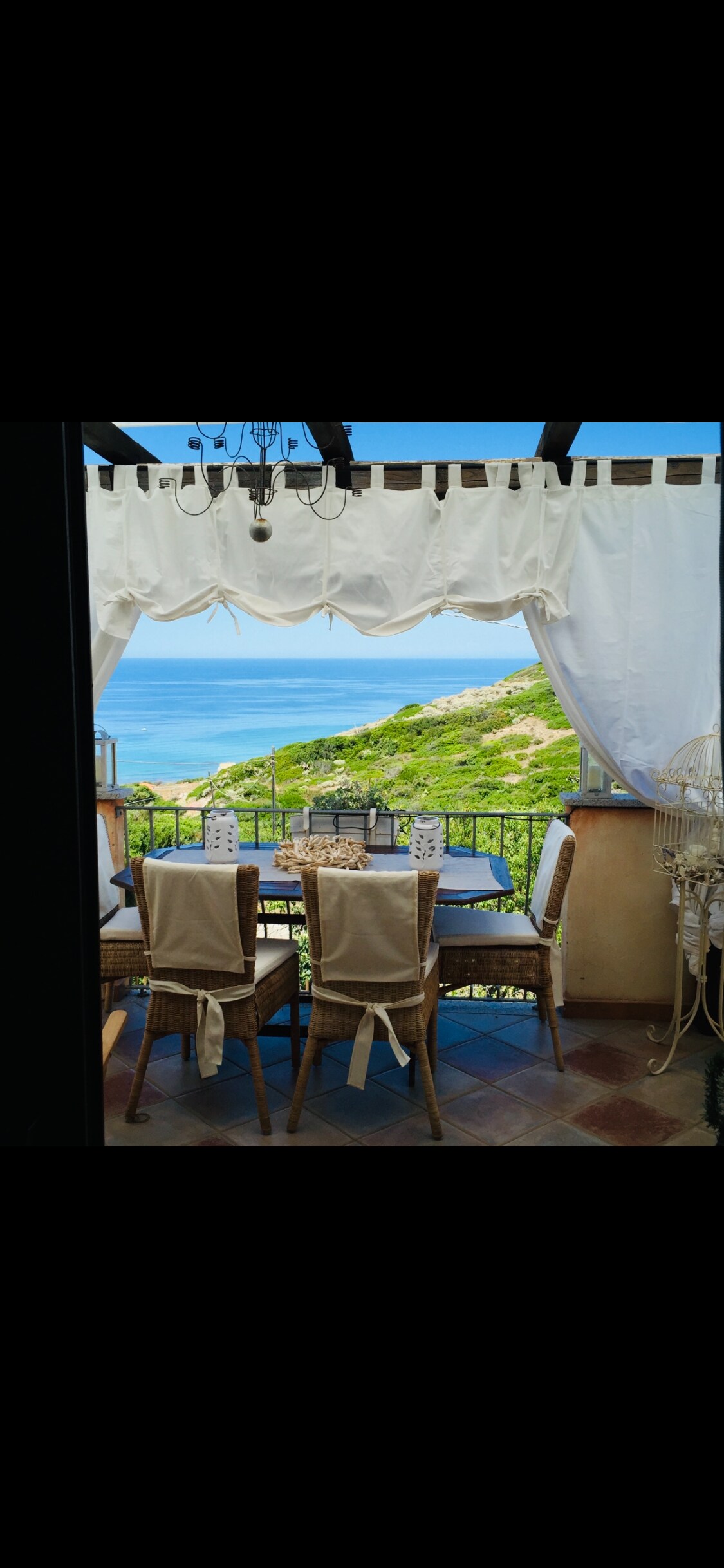 An outdoor terrace is displayed with a dining table surrounded by four chairs. Soft, flowing white curtains frame the view of lush greenery and the sea beyond. A serene atmosphere is created by the surrounding natural landscape.