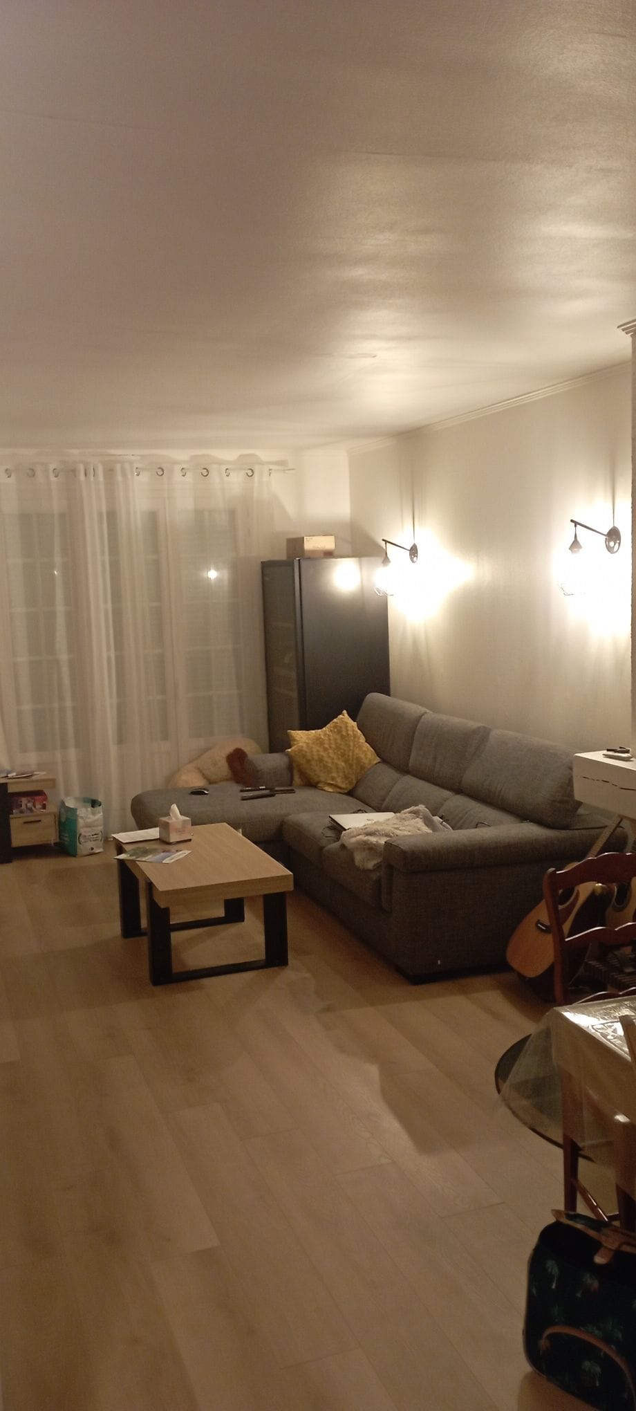 A spacious living area features a comfortable gray sofa accompanied by a yellow throw pillow. A coffee table sits in front, and warm lighting is provided by wall sconces. Large windows covered by sheer curtains allow natural light to fill the room.