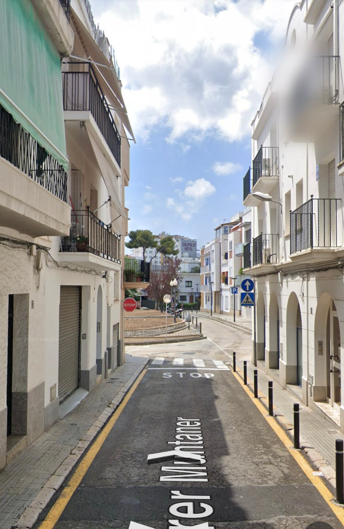A quiet street view features a narrow road lined with buildings on either side, showcasing balconies and arches. Street signs indicate traffic directions, while a few trees and clouds can be seen in the background, enhancing the serene atmosphere of the area.