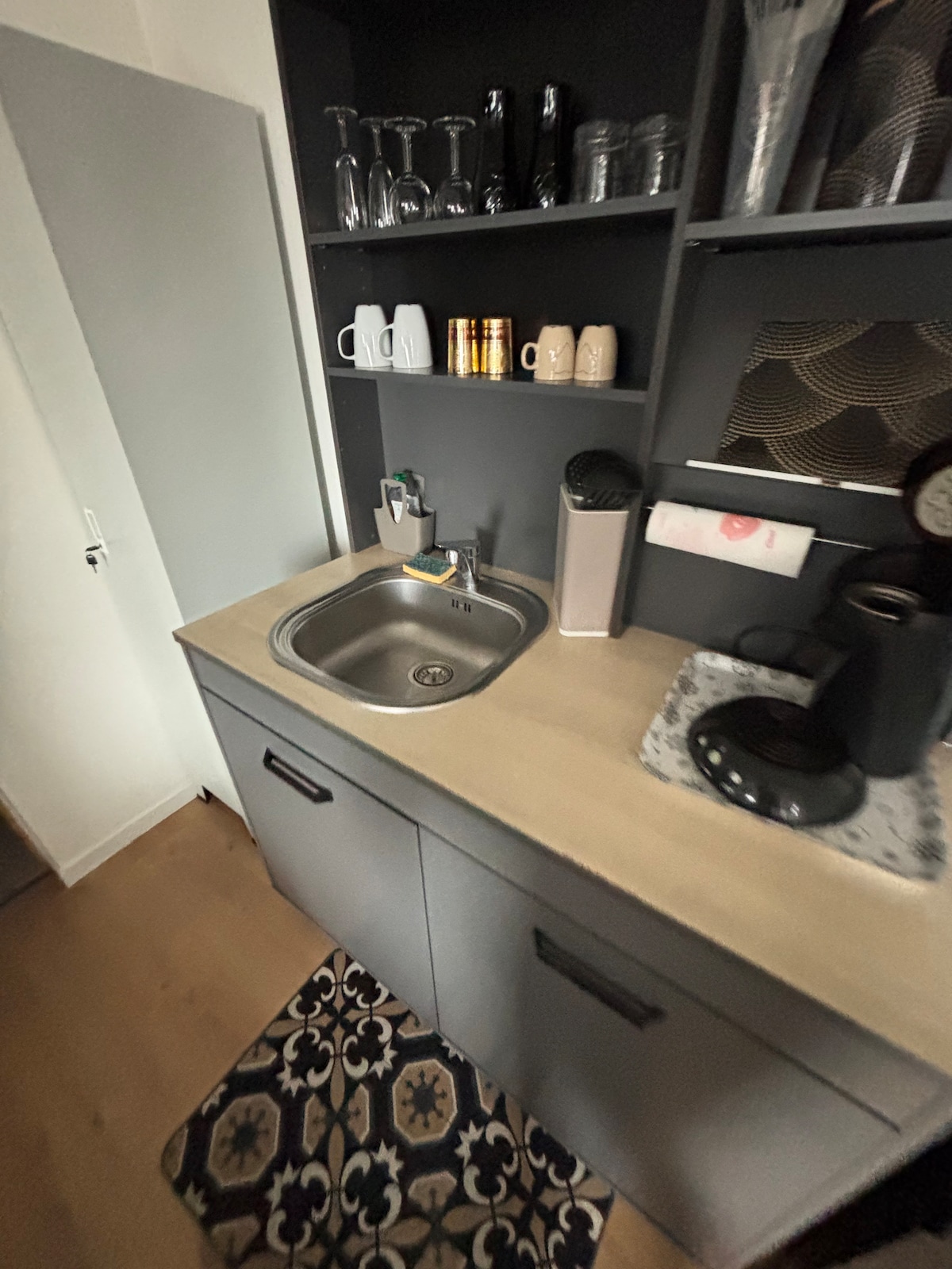 The kitchenette features dark cabinetry with an integrated sink and a coffee machine. Glasses and mugs are neatly arranged on the upper shelf, while a decorative rug adds texture to the floor. A light counter surface complements the modern appliances.
