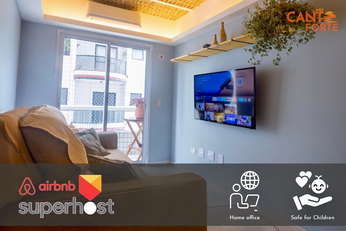 A modern living area features a comfortable sofa and a 50” Ultra HD Smart TV mounted on the wall. A small shelf adorned with greenery adds a touch of nature. Large windows provide ample natural light and access to an outdoor space.