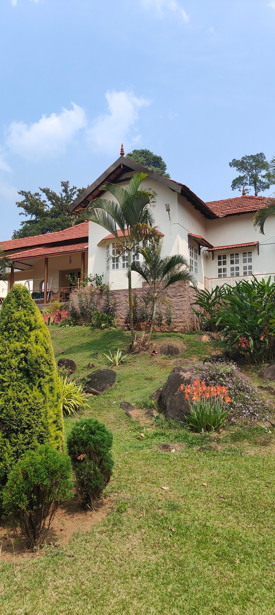 A charming bungalow is set amidst a lush green landscape, featuring manicured gardens with various plants and flowering shrubs. The white exterior, accented by a red roof, blends harmoniously with the surrounding nature. Trees and ornamental plants frame the entrance, enhancing the tranquil atmosphere.