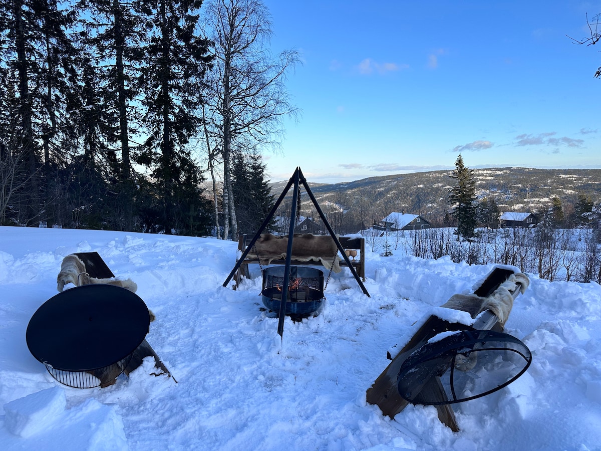 An outdoor fire pit is surrounded by wooden benches, situated on a snowy landscape. Scenic views of distant mountains and trees provide a tranquil backdrop. The fire pit features a grill grate, ready for cooking. The open sky allows for natural light and a serene ambiance.